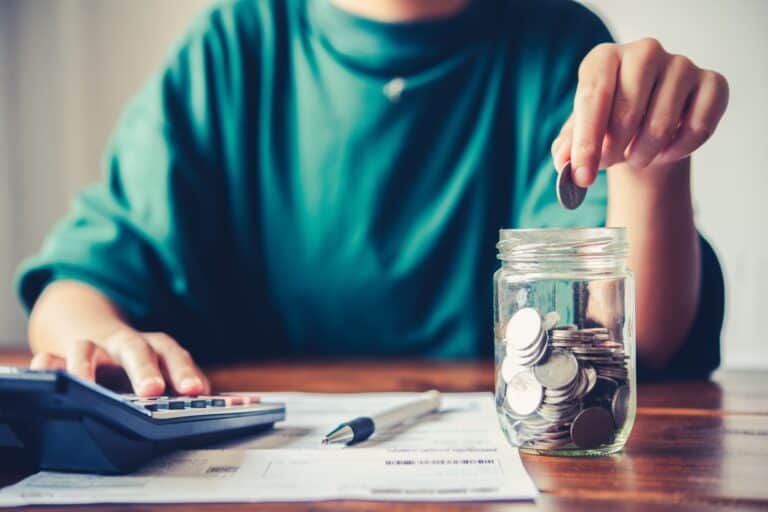 A woman is putting a coin into a jar with a pen, a document, and a calculator on the table in the concept of saving money for the move to Wandsworth.